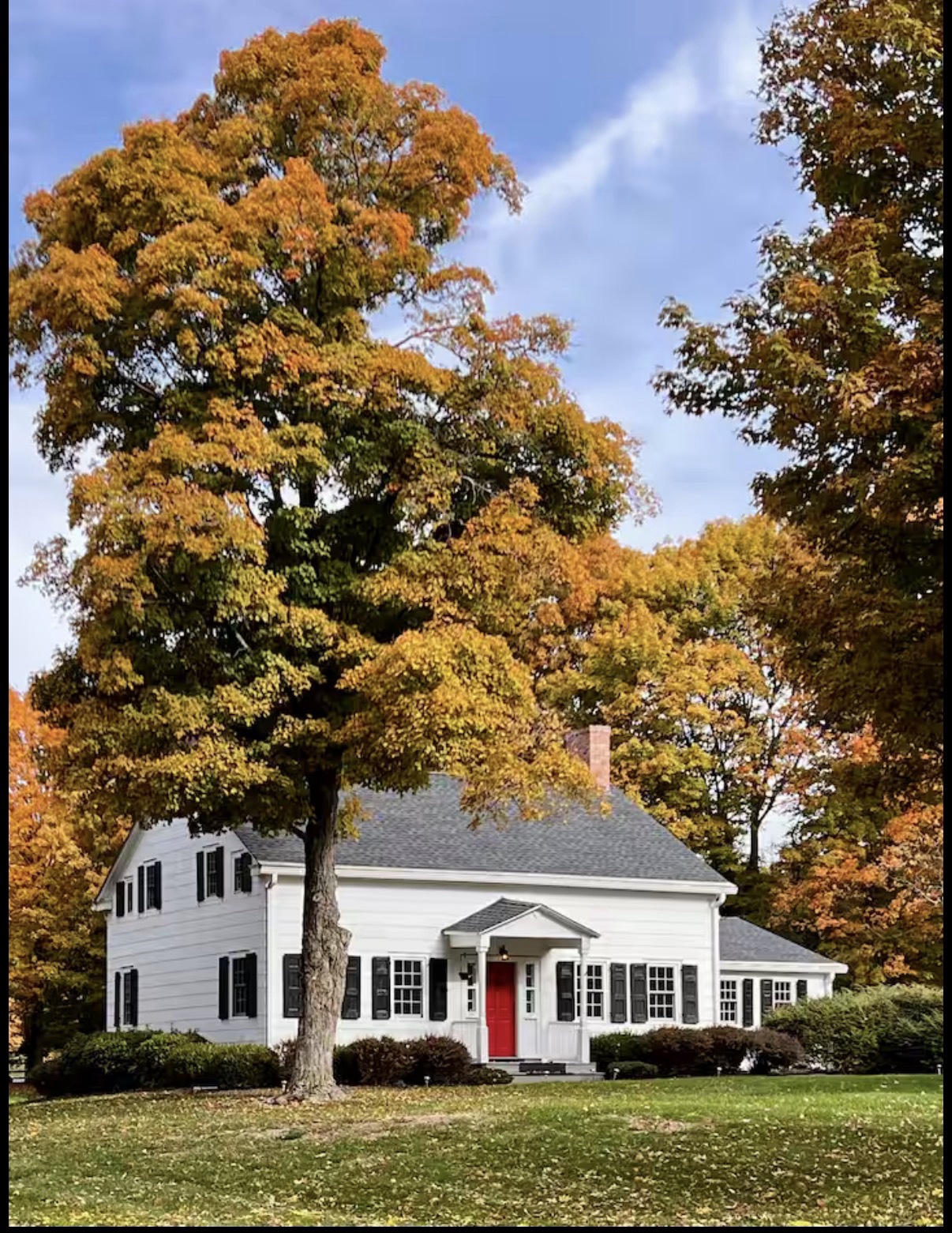 Front of the white farmhouse with its red door beneath vivid fall foliage