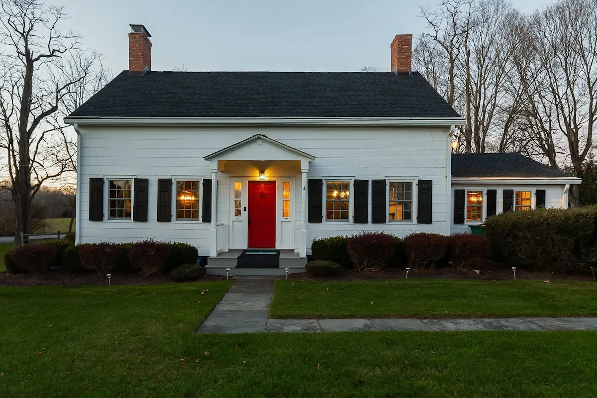 Front of the white farmhouse with its red door at dusk